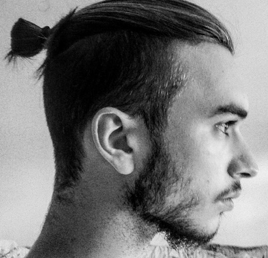 A black and white photograph of a hipster guy with a manbun undercut hairstyle and some stubble on his face