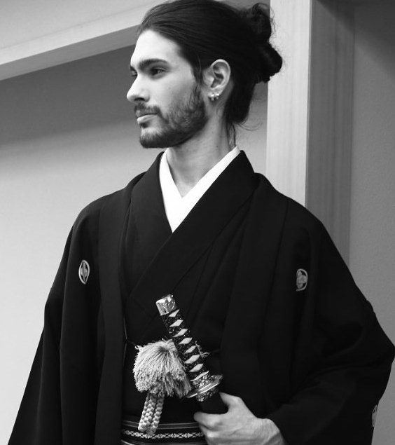 A photograph of a white Samurai male with a cool Chonmage hairstyle which is also known as a topknot hairstyle for his long dark hair as he stands up with his sword ready for fighting