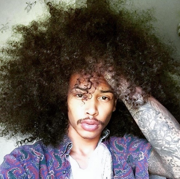 A photograph of a black hipster male with his very-long kinky curly hair worn as an afro hairstyle and combined with a thin mustache on his upper lip
