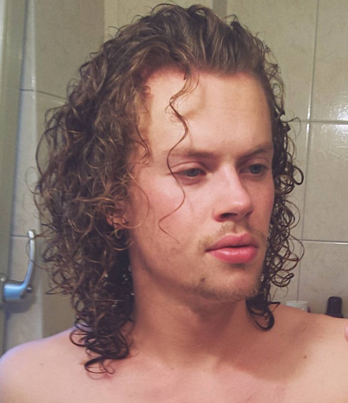 A photograph of a handsome white male with long curly hair that has been slicked back with a good hairstyling gel