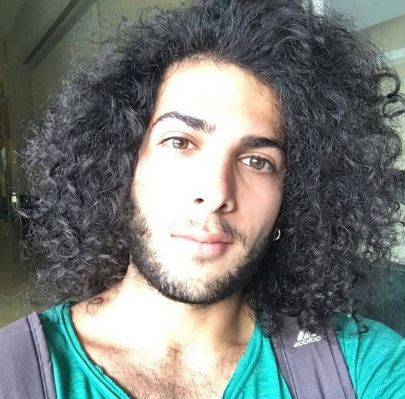 A photograph of an Arab male with long curly hair combed in a side part hairstyle and cut in layers