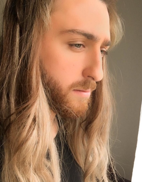 A side-view picture of a handsome blonde guy with long wavy hair that he has styled with hair gel and a blow dryer