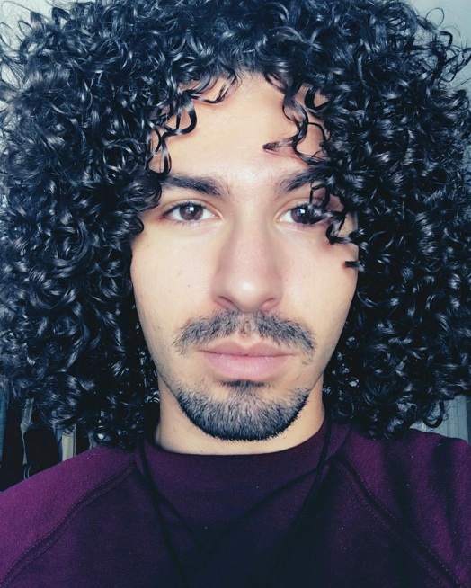 A photograph of a Latin male from New York who has his long curly hair styled with a layered haircut which makes his coils look similar to the ones of Rogelio Samson