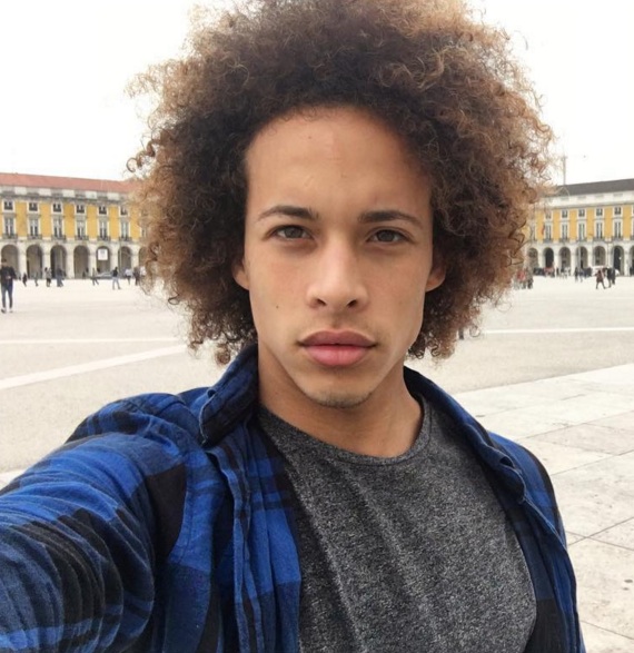 A photograph of a mulatto French male with blonde kinky curly hair that is at a current long length of ten inches but which is still puffing out