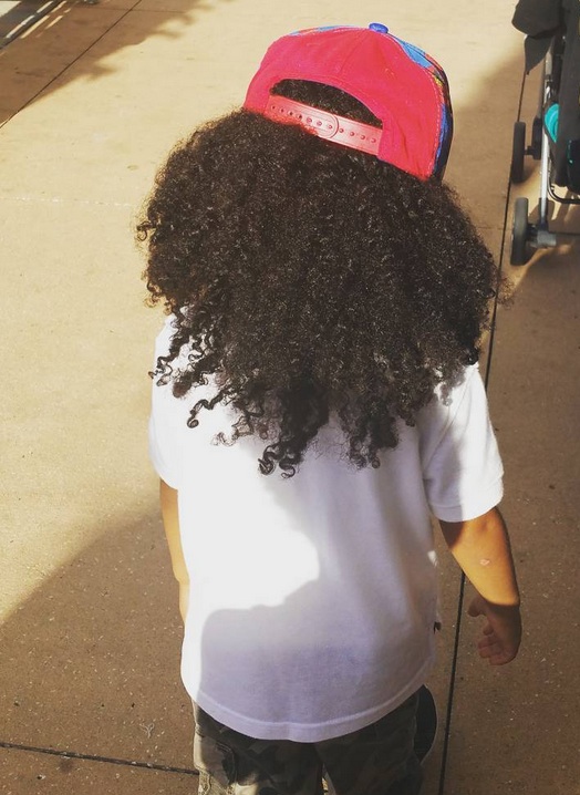 A photograph of an African-American toddler with long kinky curly hair styled back under a baseball cap