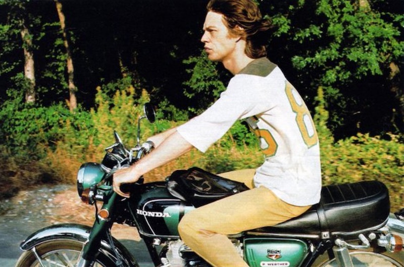 A high-quality photo of Mick Jagger with long hair riding a Honda motorcycle at high speed with no protective gear