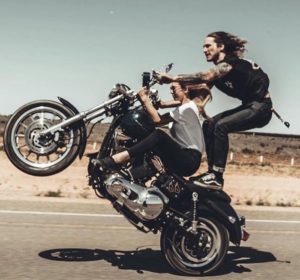 A photo of a biker couple in which the long haired male is riding the motorbike dangerously with no motorcycle gear